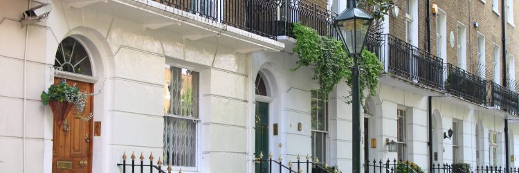 Row of elegant cream-colored Georgian townhouses with black iron railings, arched ground-floor windows, and neatly trimmed potted plants on balconies.