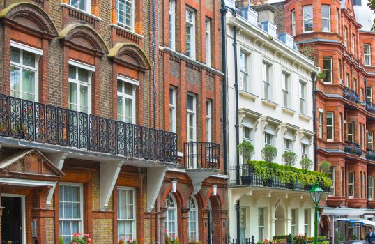 Colorful row of historic brick buildings with decorative balconies, white-framed windows, and lush greenery lining the facades on a narrow street.