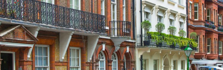 Colorful row of historic brick buildings with decorative balconies, white-framed windows, and lush greenery lining the facades on a narrow street.